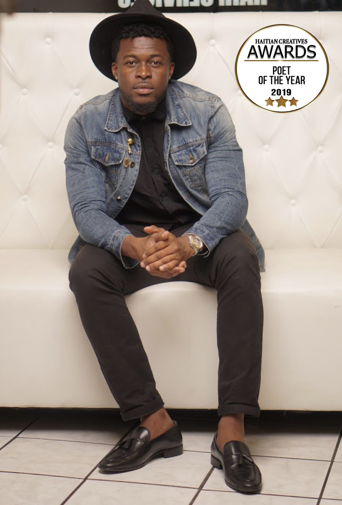 Man of african decent seated on white seat, with ablack hat, denim jacket and grey dress pants. Black dress shoes