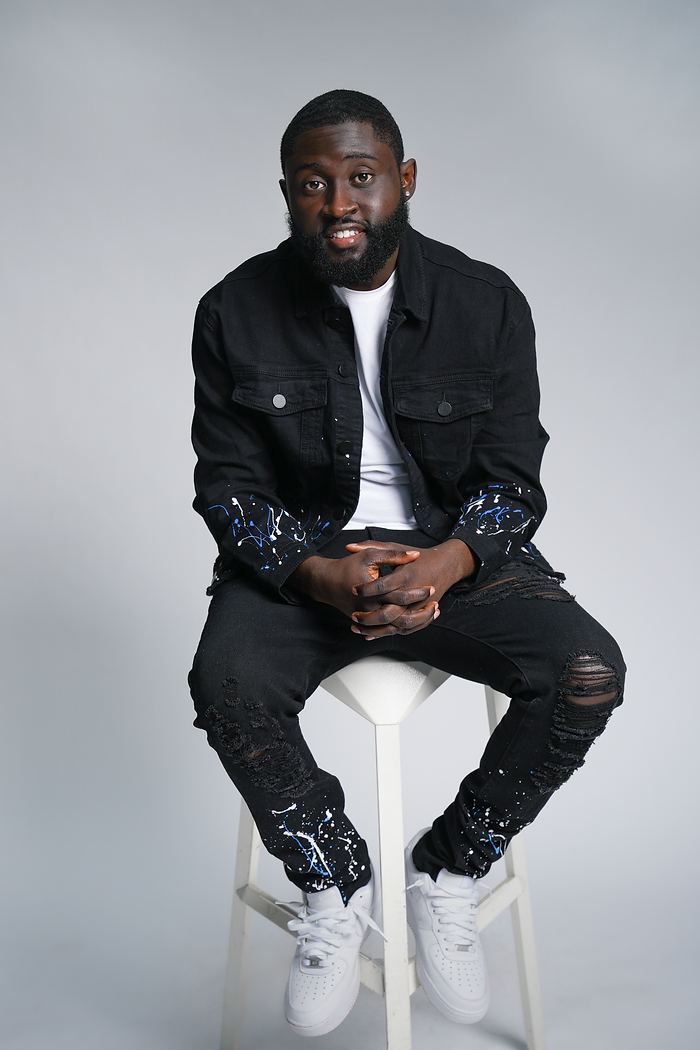 BLACK man seated on a white stool. weating a black denim outfit with a white shirt.