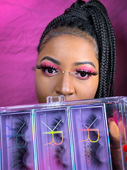 Image of woman with african decent with imae of eyes with dramatic pink eye shadow, lashed, and braided ponytail. A pack of 3 lashes cover her mouth as she displays them.