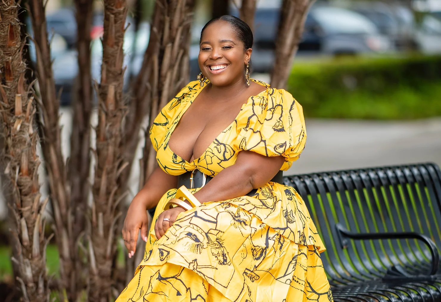 woman of african decent dressed in long yellow dress with hair pulled back tight, smile forward while sitting on the edge of a green park bench.