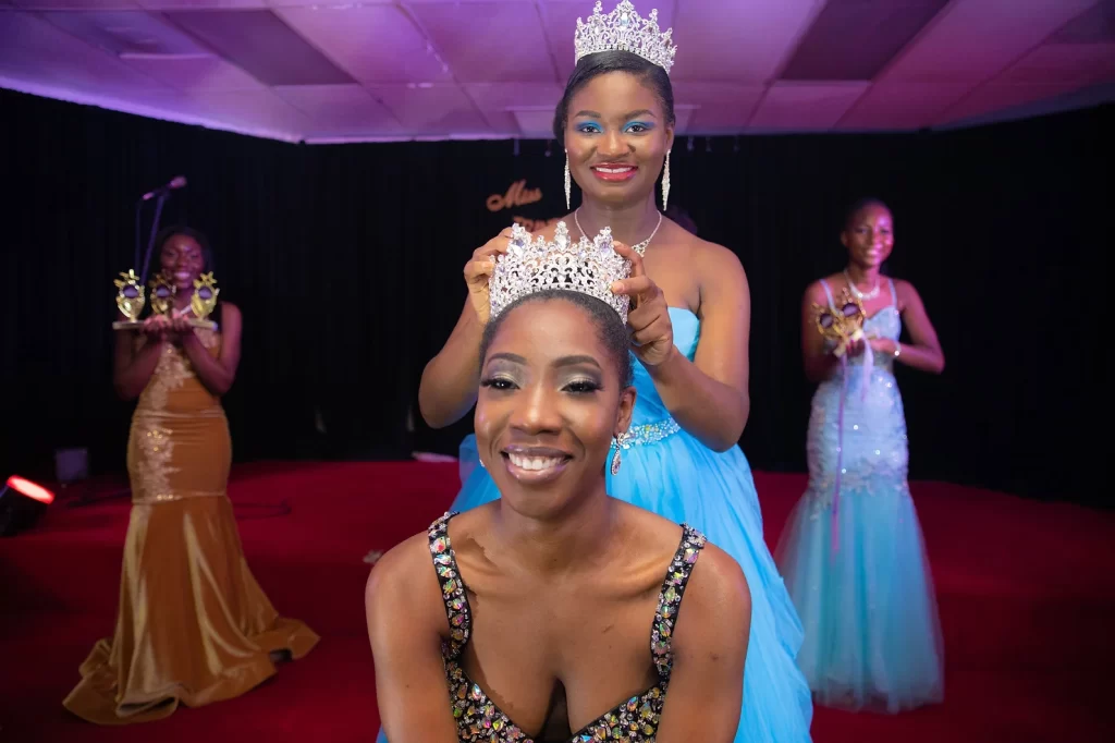 in the center there is a woman of african decent, that is being crowned by a woman in blue gown. while two other woman of african decent stand in the back with aawards. 