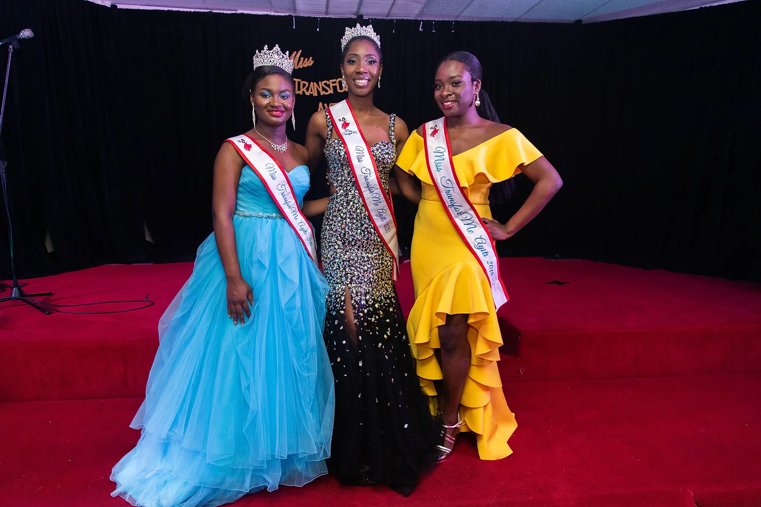 3 women of african decent wearing pagent sashes acccross ther bodies from right to left they are wearing pagent dress. The woman on the right is wearing a gold gown, the woman on the far left blue dress, the woman in the center is wearing a black dress.