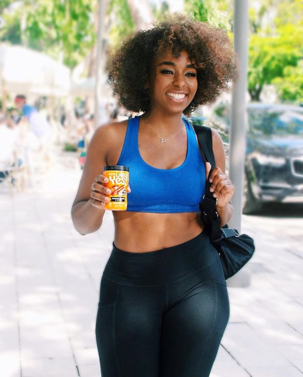 woman with african decent wearing blue sports bra, and black leggings. She is holding a beverage in the right hand and hand bag in the left hand.