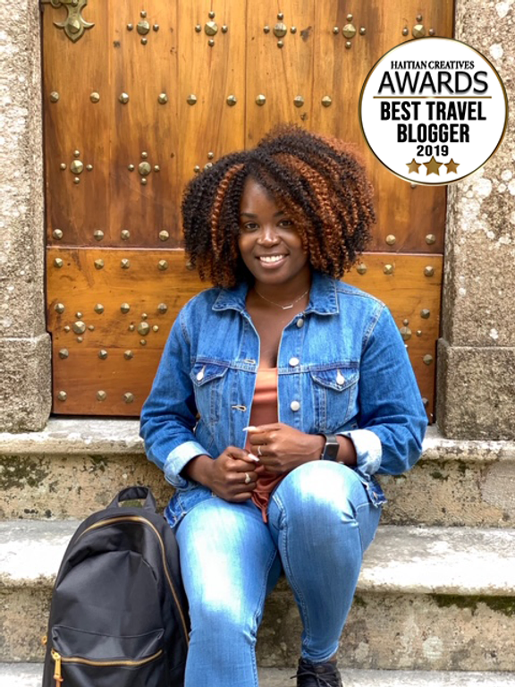 woman of african decent, is sitting outside in a denim button up and jeans. Mixed brown afro and a smile.Bag on her left side of cement steps. 